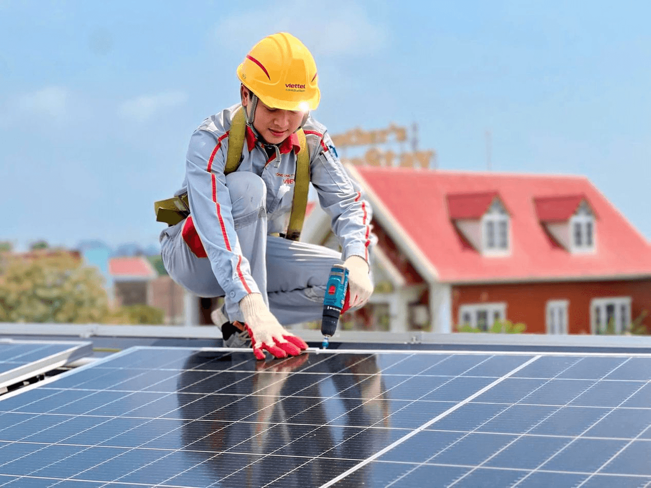 Worker in yellow helmet and grey uniform installing solar panel with drill on rooftop